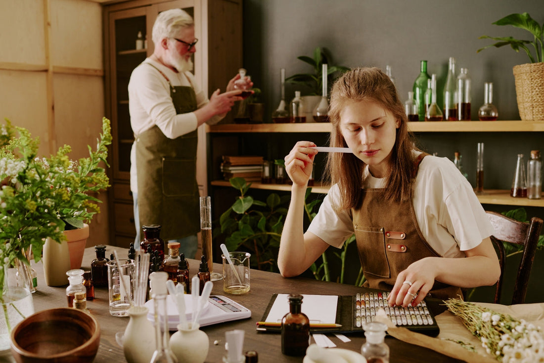 Two perfumers working in a fragrance studio, with one smelling a scent strip at a table filled with perfume bottles, droppers, and fresh botanicals