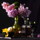 Still life with pink hydrangeas, lemons, a glass perfume-style decanter, and a green goblet arranged on a dark table against a moody background