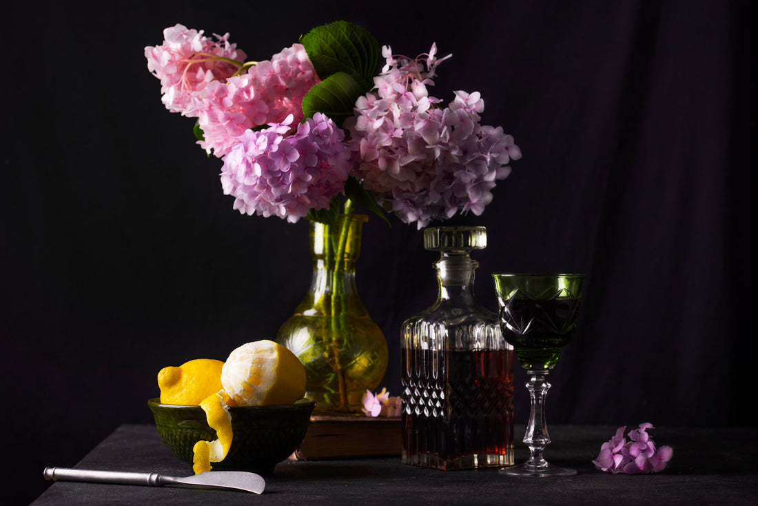 Still life with pink hydrangeas, lemons, a glass perfume-style decanter, and a green goblet arranged on a dark table against a moody background